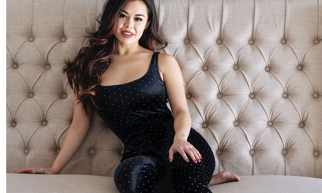 A beautiful brunette model smiling sitting on a. fabric tufted beige linen couch wearing a black velvet loungewear set by the brand vanity couture