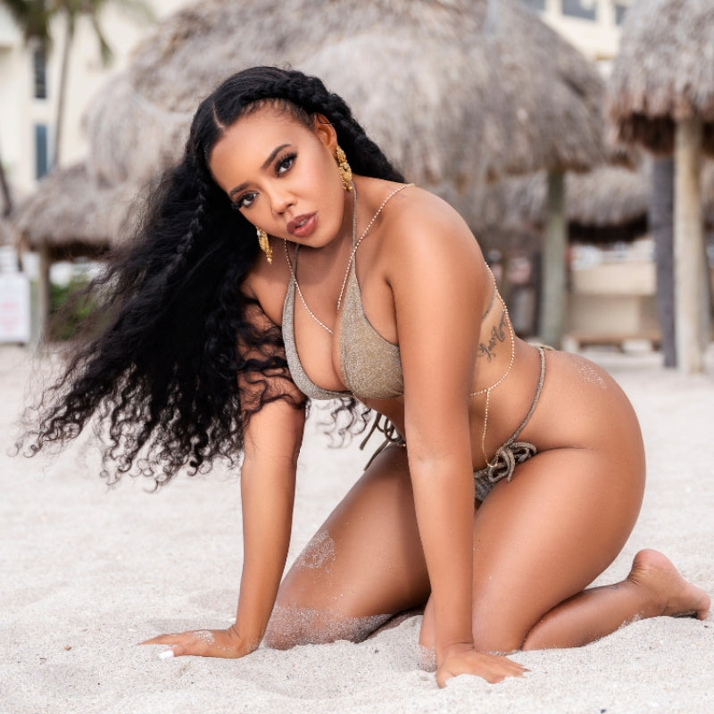 Model And fashion designer Angela Simmons kneeling down in sand on beach posing looking at the camera wearing a sparkly gold Brazilian bikini set from her collection with vanity couture